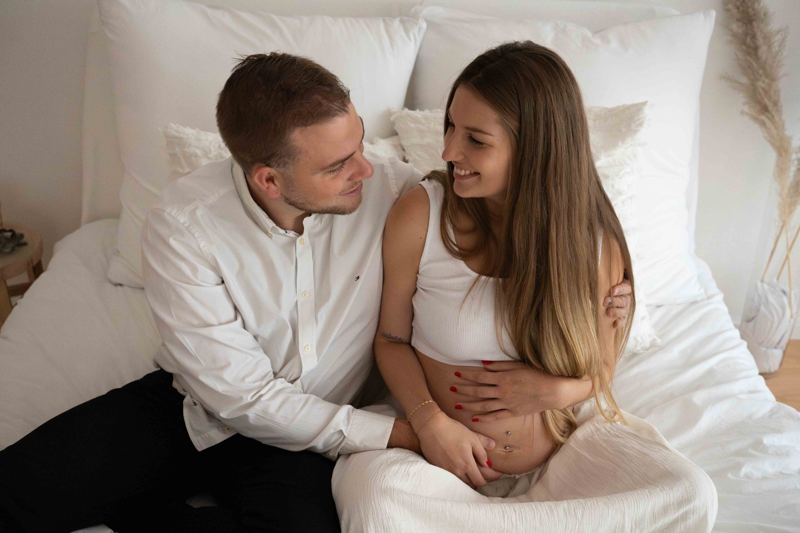 Schwangere-Frau-und-Partner-sitzen-beim-Babybauchshooting-auf-Bett-und-schauen-sich-an